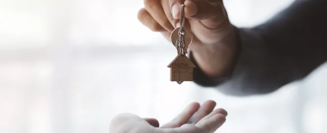 Hand holding house-shaped keychain labeled "Home" handing it to another hand