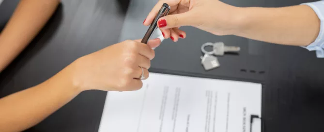Two hands exchange a pen over a rental contract on a black table, with keys and takeaway cups nearby—suggesting the signing of a lease agreement.