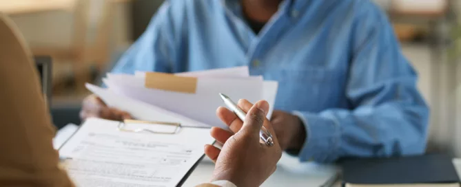 Person in blue shirt holding papers while another hand holds a pen over a clipboard with documents, illustrating a professional meeting about lease violations.
