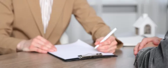 Person in beige blazer signing documents at a table with keys and a house model, illustrating safe handling of tenant complaints in Dallas–Fort Worth.